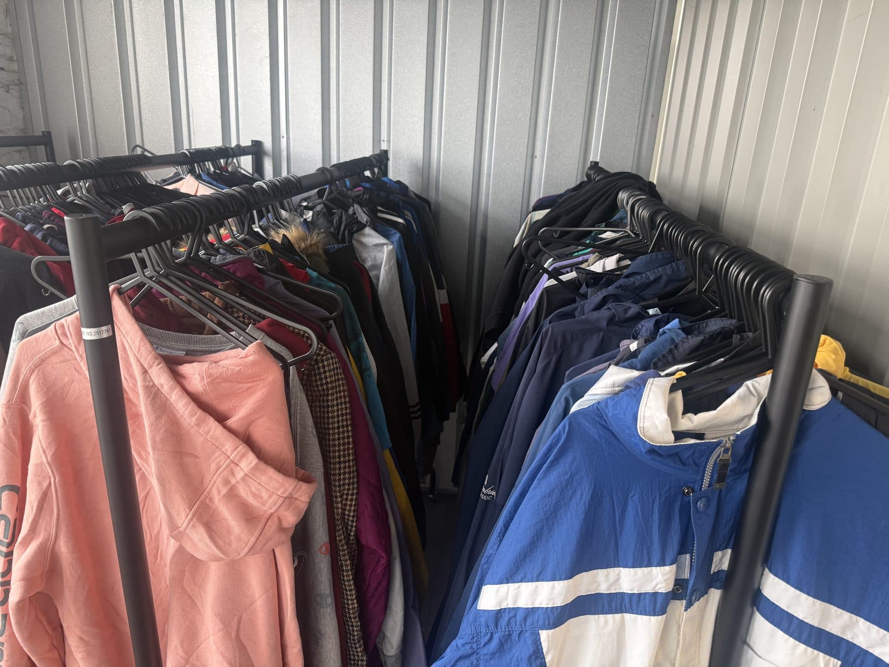 Two rails of vintage garments on black hangers, including a coral hooded sweatshirt, a checked shirt and blue and white windbreakers.